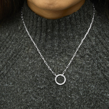 Close-up front view of a model wearing a white metal necklace with a circular pendant featuring pave-set white gemstones, shown against a dark textured top.