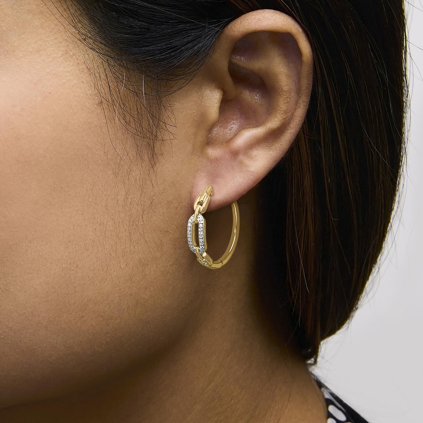 Close-up side view of a model wearing a yellow metal hoop earring with an elongated oval link adorned with pave-set white gemstones.