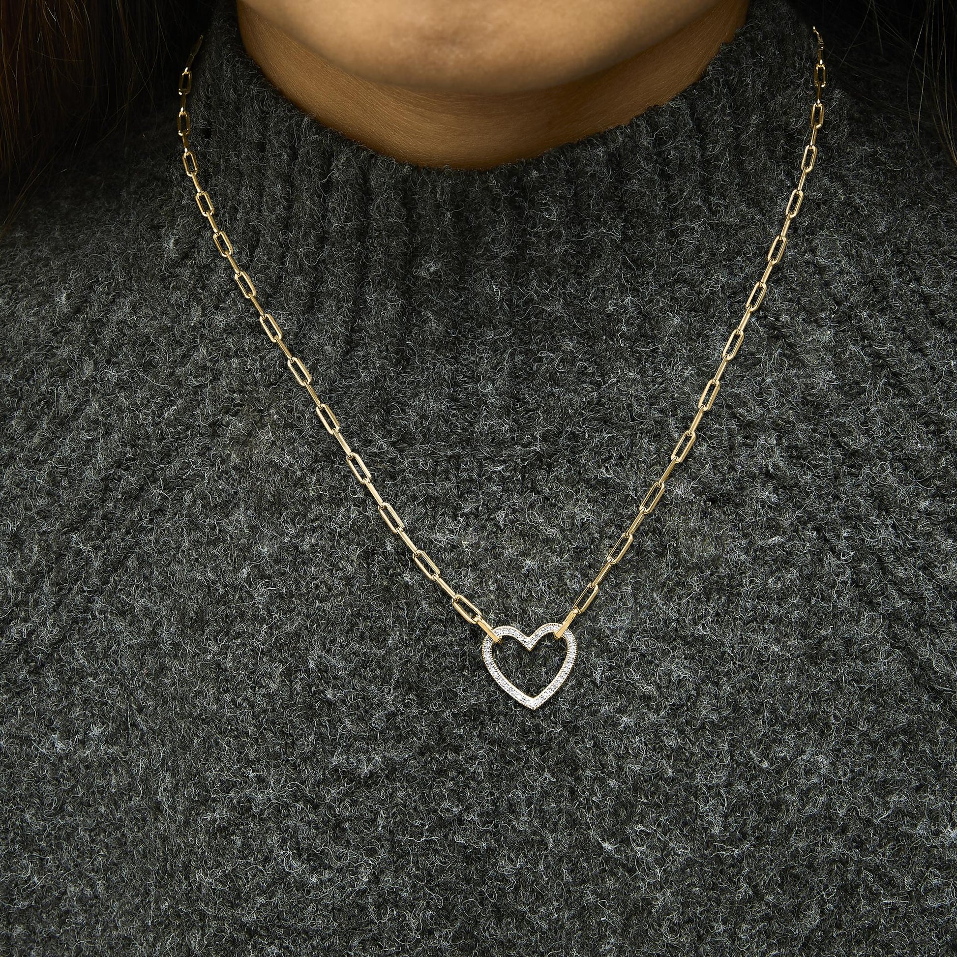 Close-up front view of a model wearing a yellow necklace with a heart-shaped pendant featuring white gemstones in a pave setting, on a textured dark gray sweater.