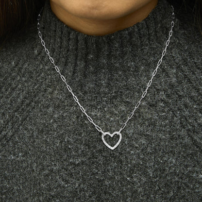 Close-up front view of a model wearing a white metal necklace with a heart-shaped pendant featuring pave-set round white gemstones, shown against a dark textured sweater.