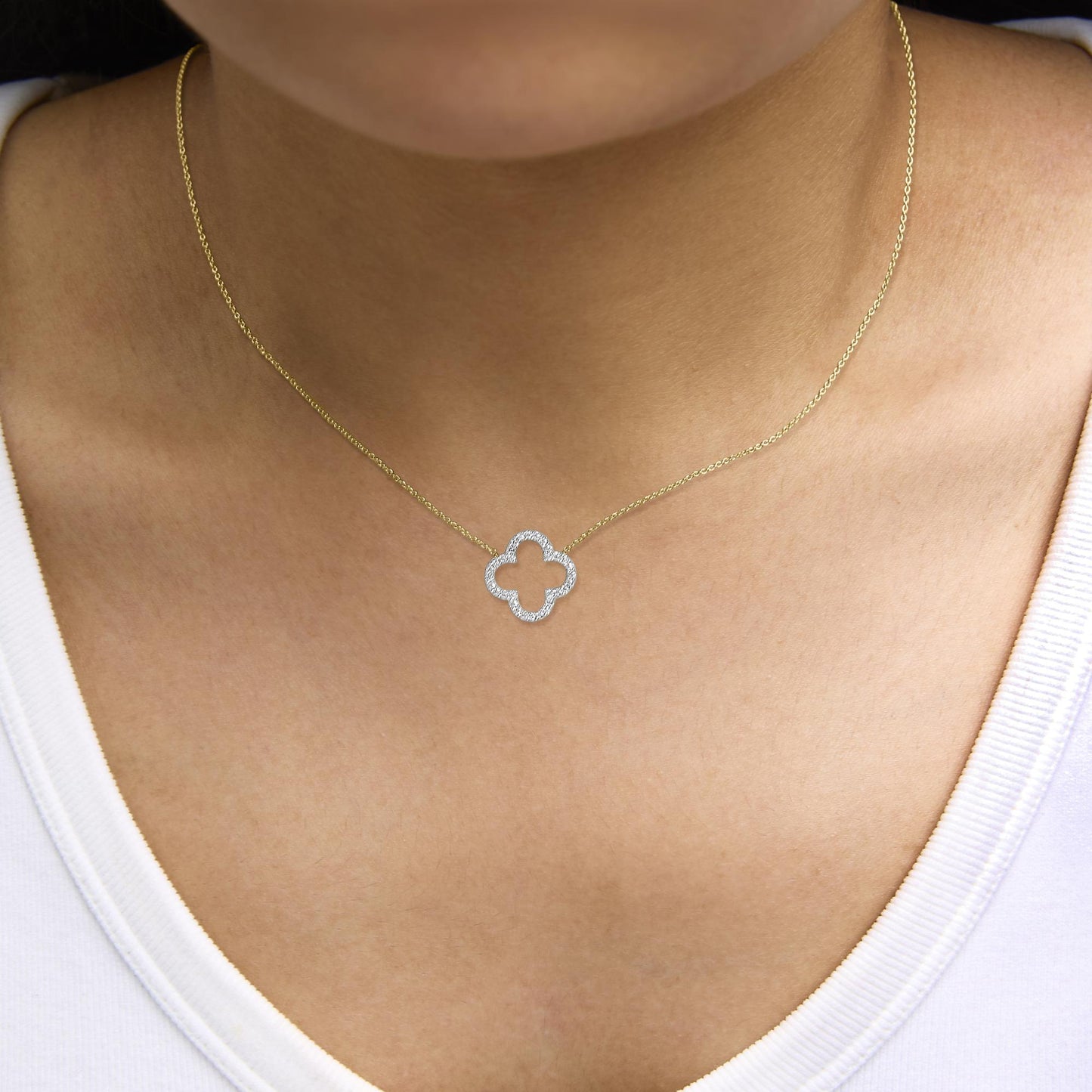 Yellow metal necklace with a quatrefoil pendant encrusted with round white gemstones in prong settings, shown worn on a model, close-up front angle.