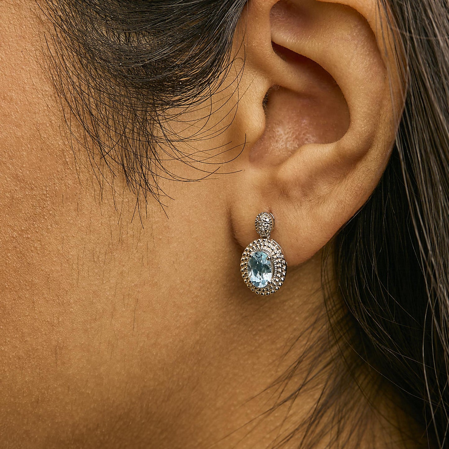 Close-up side view of white metal earrings shown worn on a model’s ear, featuring an oval blue gemstone surrounded by small white gemstones in a pave setting.