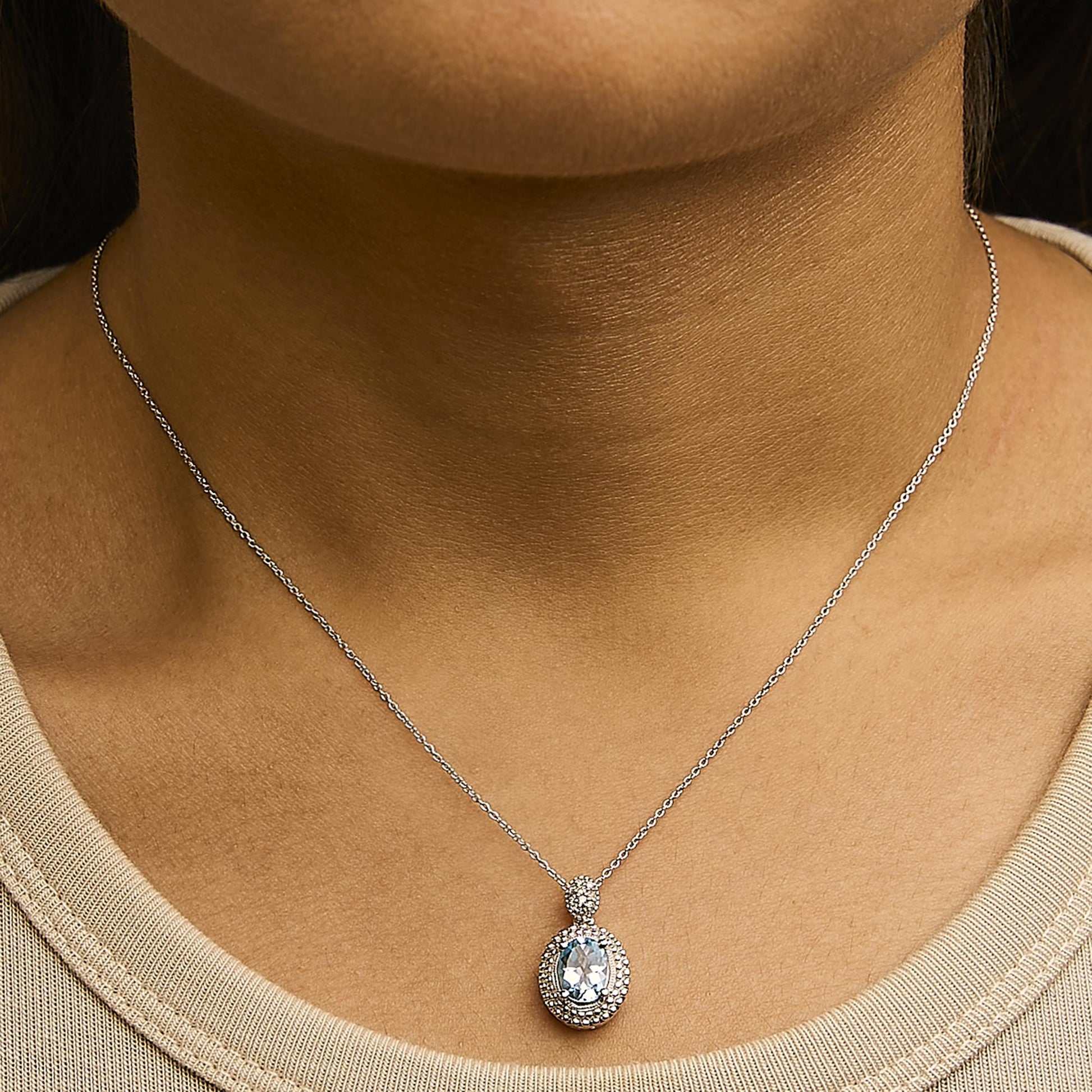 Close-up front view of a white necklace with a pendant featuring an oval blue gemstone surrounded by small white gemstones, worn on a model.