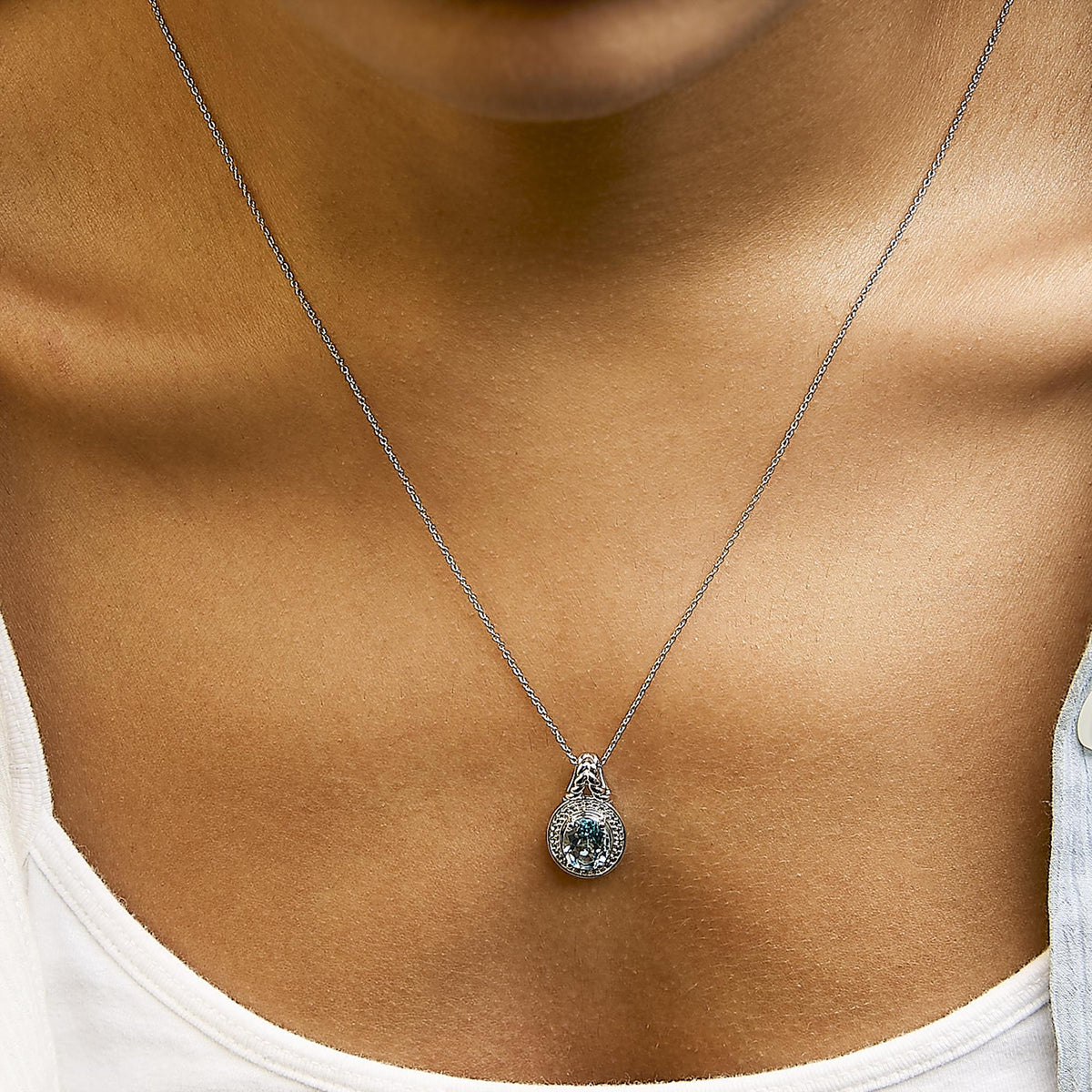 Close-up front view of necklace with a white metal chain and a pendant featuring an oval blue gemstone surrounded by round white gemstones in prong settings, shown worn on a model.