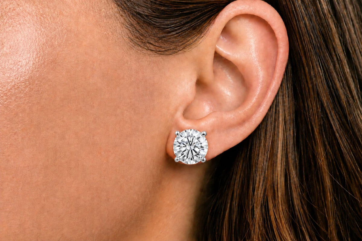 Close-up side view of a model's ear wearing a white metal stud earring with a round white gemstone in a four-prong setting.