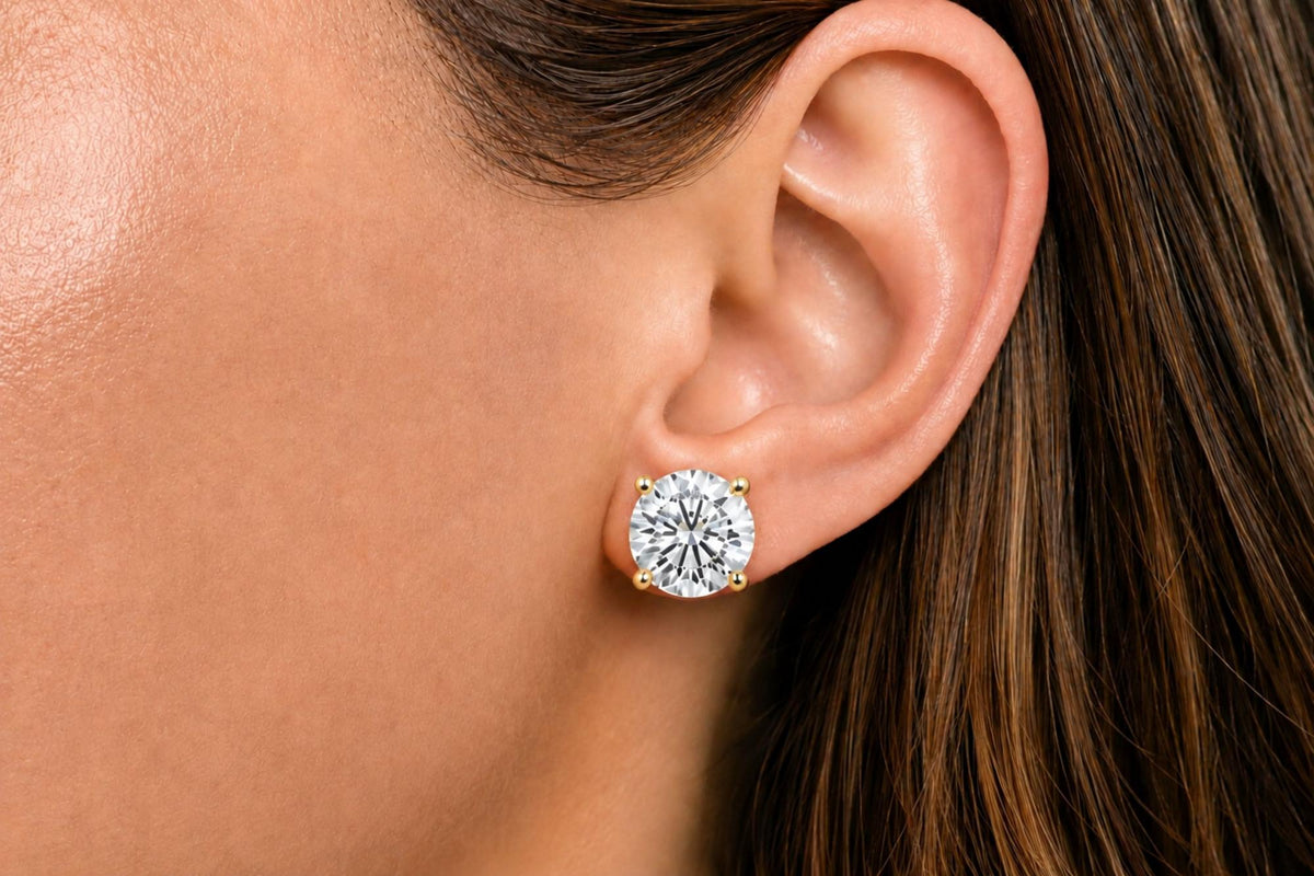 Close-up side view of a model's ear wearing a yellow metal stud earring with a round white gemstone in a four-prong setting.