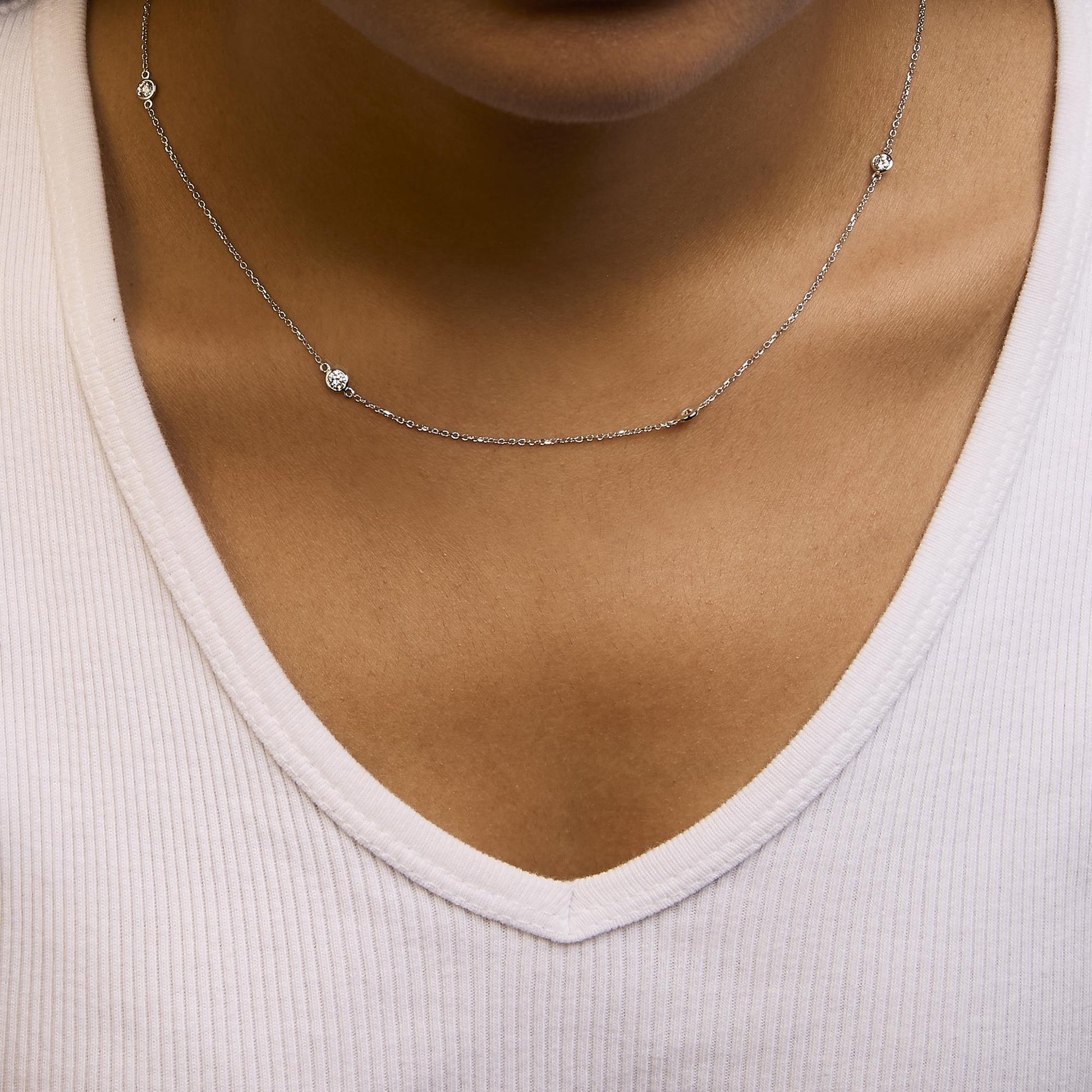 Close-up front view of a white metal necklace with five round bezel-set white gemstones, worn on a model with a white ribbed v-neck top.