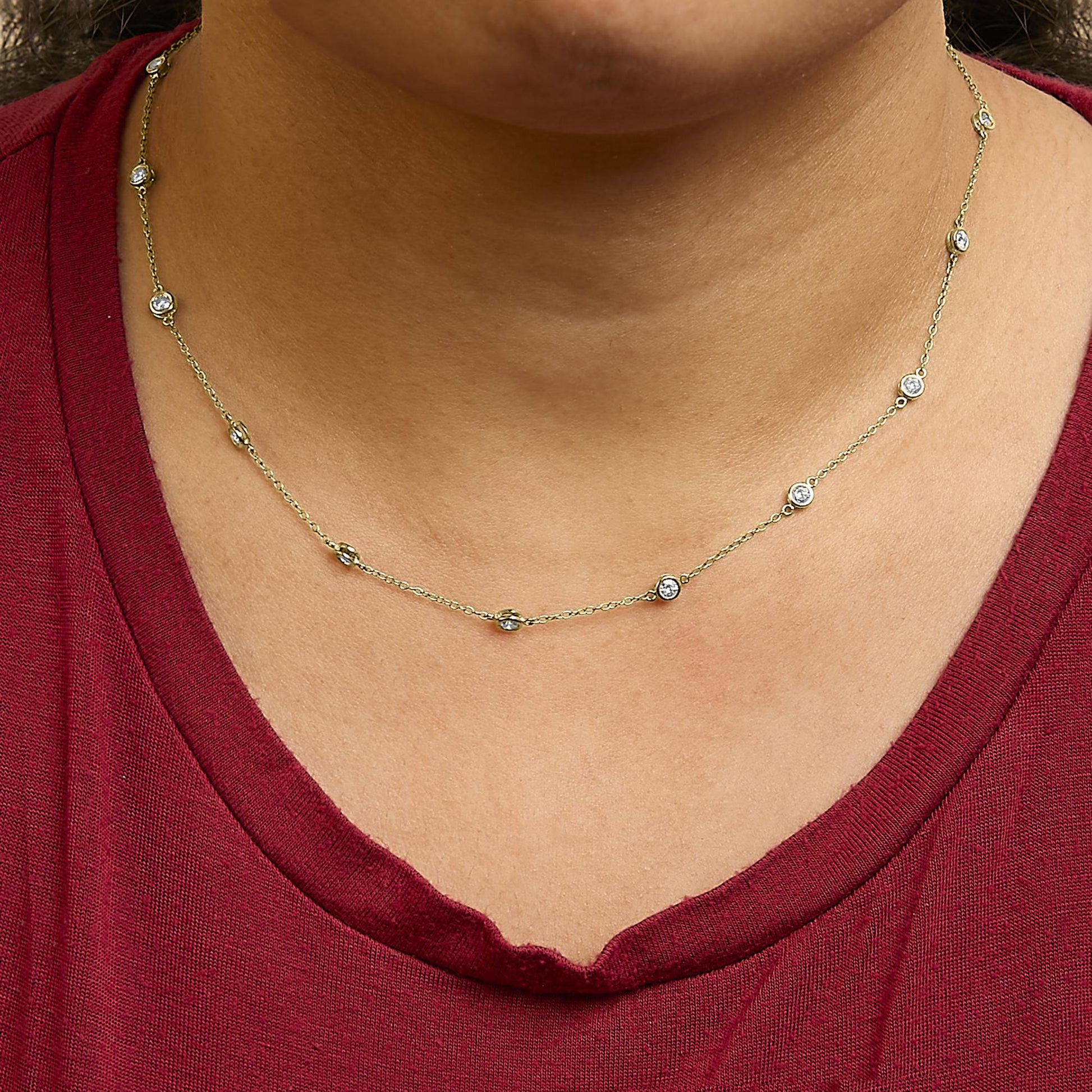 Close-up front view of yellow metal necklace with round white gemstones in bezel settings, worn on a model with a red top.