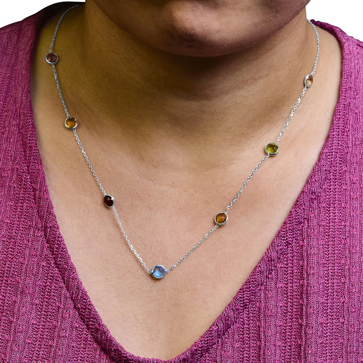Close-up front view of a white metal necklace with round bezel-set blue, purple, red, white, and smoky gemstones, worn on a model with a pink textured top.