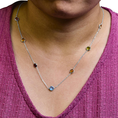 Close-up front view of a white metal necklace with round bezel-set blue, purple, red, white, and smoky gemstones, worn on a model with a pink textured top.