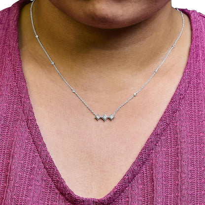 Close-up front view of a white metal necklace on a model, featuring a central section with three bezel-set princess-shaped white gemstones and a fine chain adorned with six bezel-set round white gemstones spaced along its length.