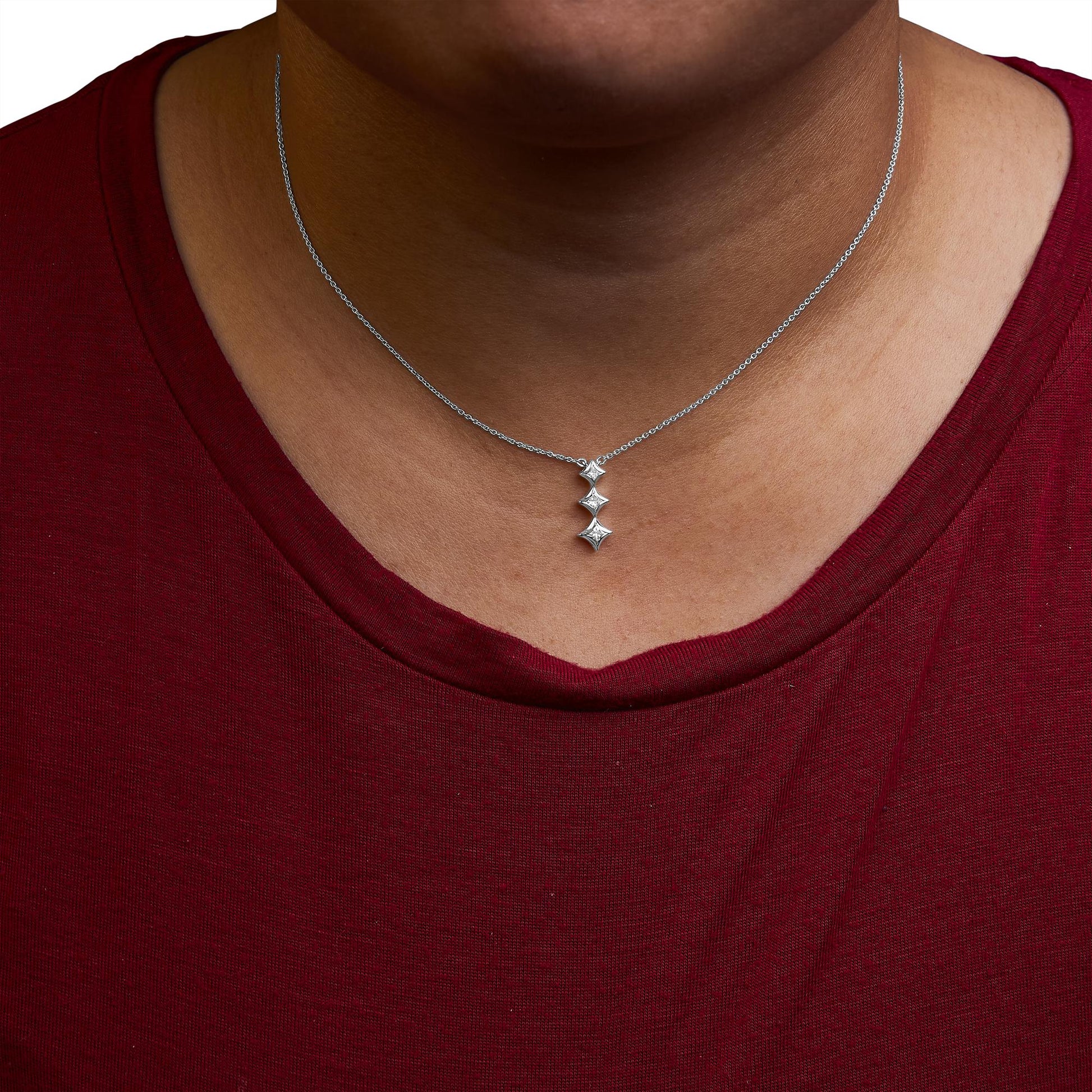 Close-up front view of a white metal necklace on a model wearing a red top, featuring a vertical pendant with three princess-shaped white gemstones in bezel settings.