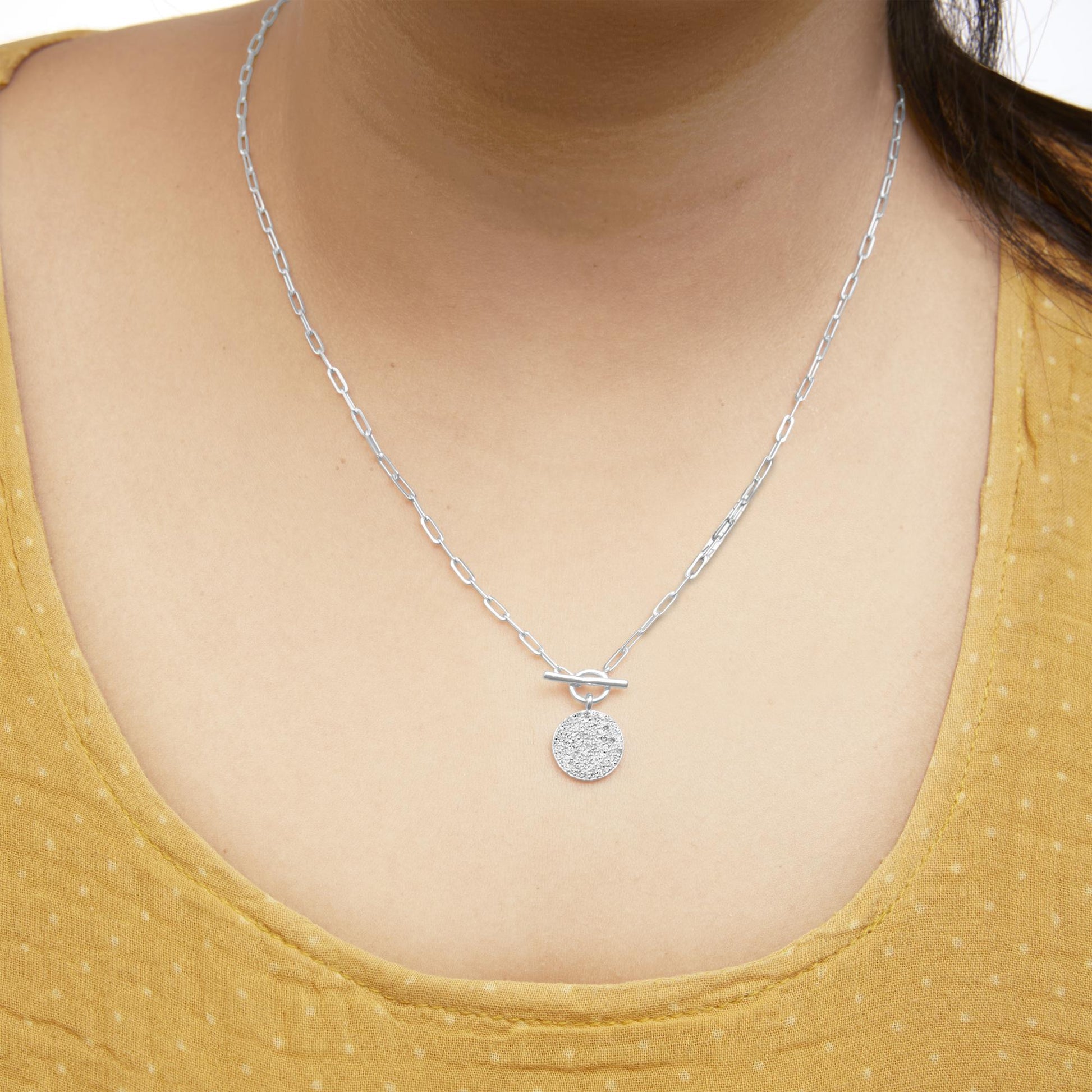Close-up front view of a white metal necklace with a chain link design and a round pendant encrusted with small white gemstones, worn on a model.