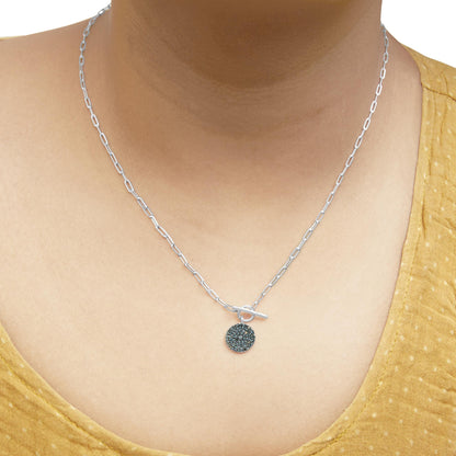 Close-up front view of a white metal necklace on a model, featuring a circular pendant encrusted with blue round gemstones in prong settings and a toggle clasp.
