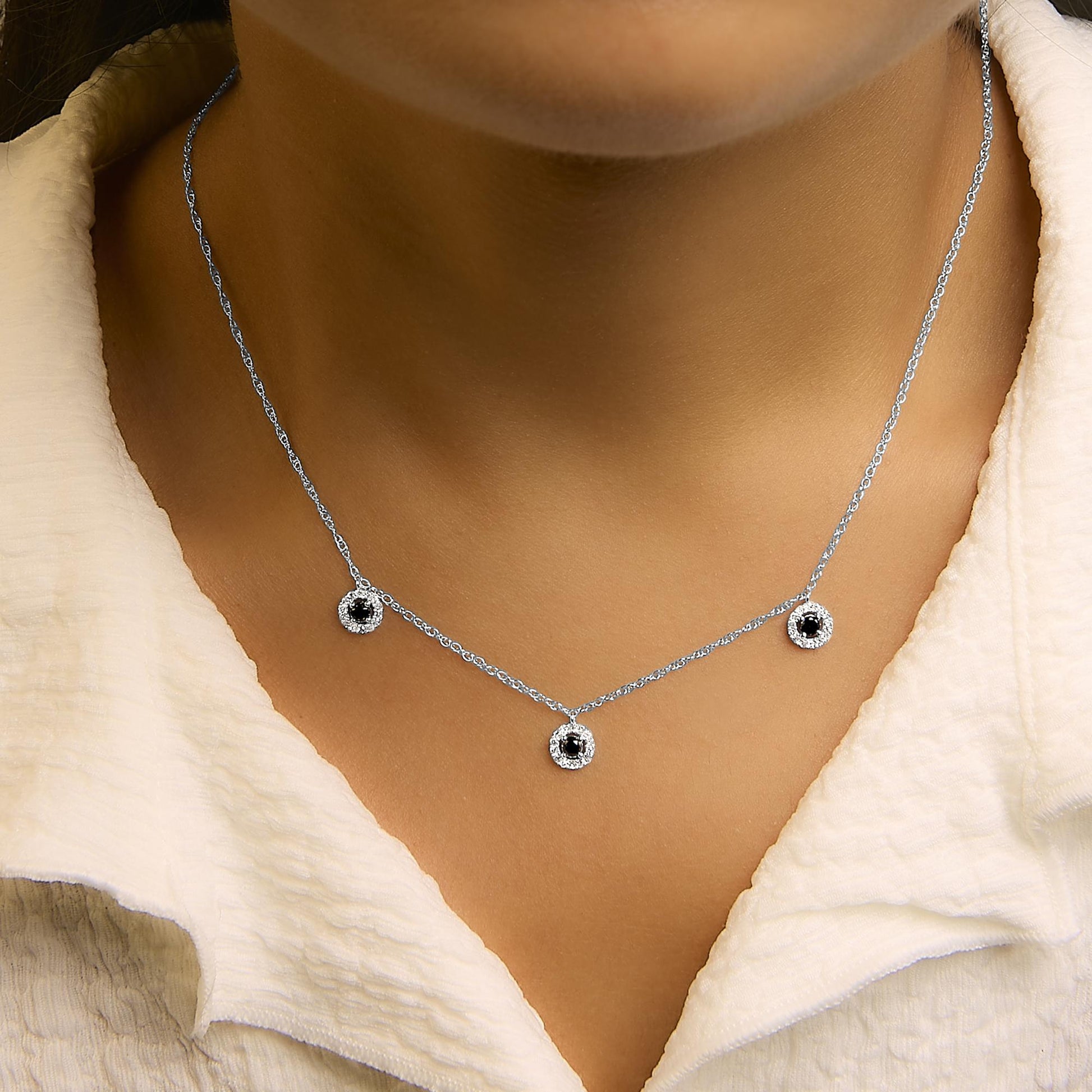 Close-up front view of a model wearing a white metal necklace with a delicate chain and three round pendants. Each pendant features a central black gemstone surrounded by small white gemstones in prong settings.