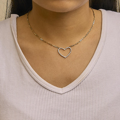 Close-up front view of a yellow metal necklace on a model, featuring a heart-shaped pendant adorned with prong-set round white gemstones, and a linked chain.