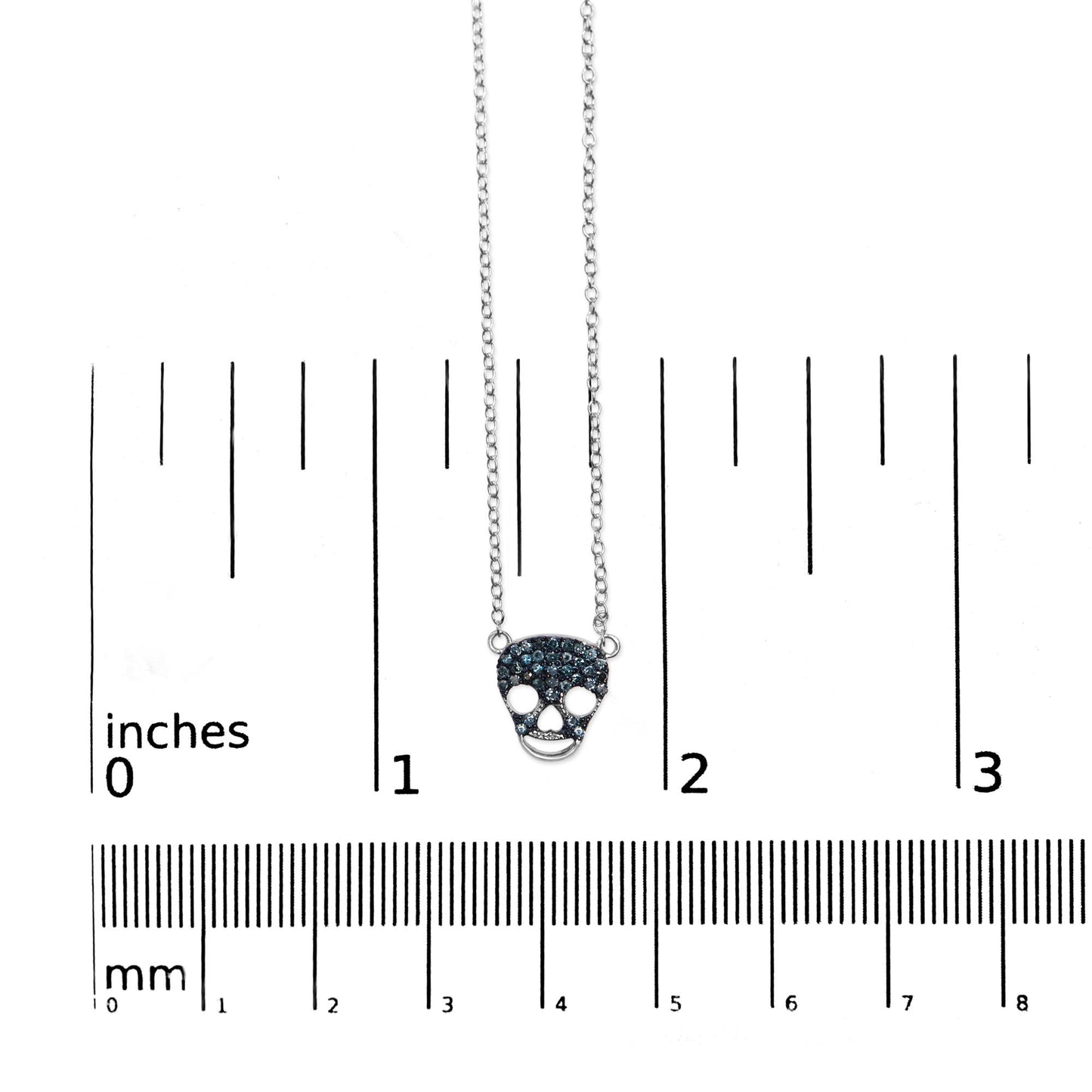 White metal necklace with skull-shaped pendant featuring round blue gemstones in prong settings, photographed from above on a ruler for size reference.