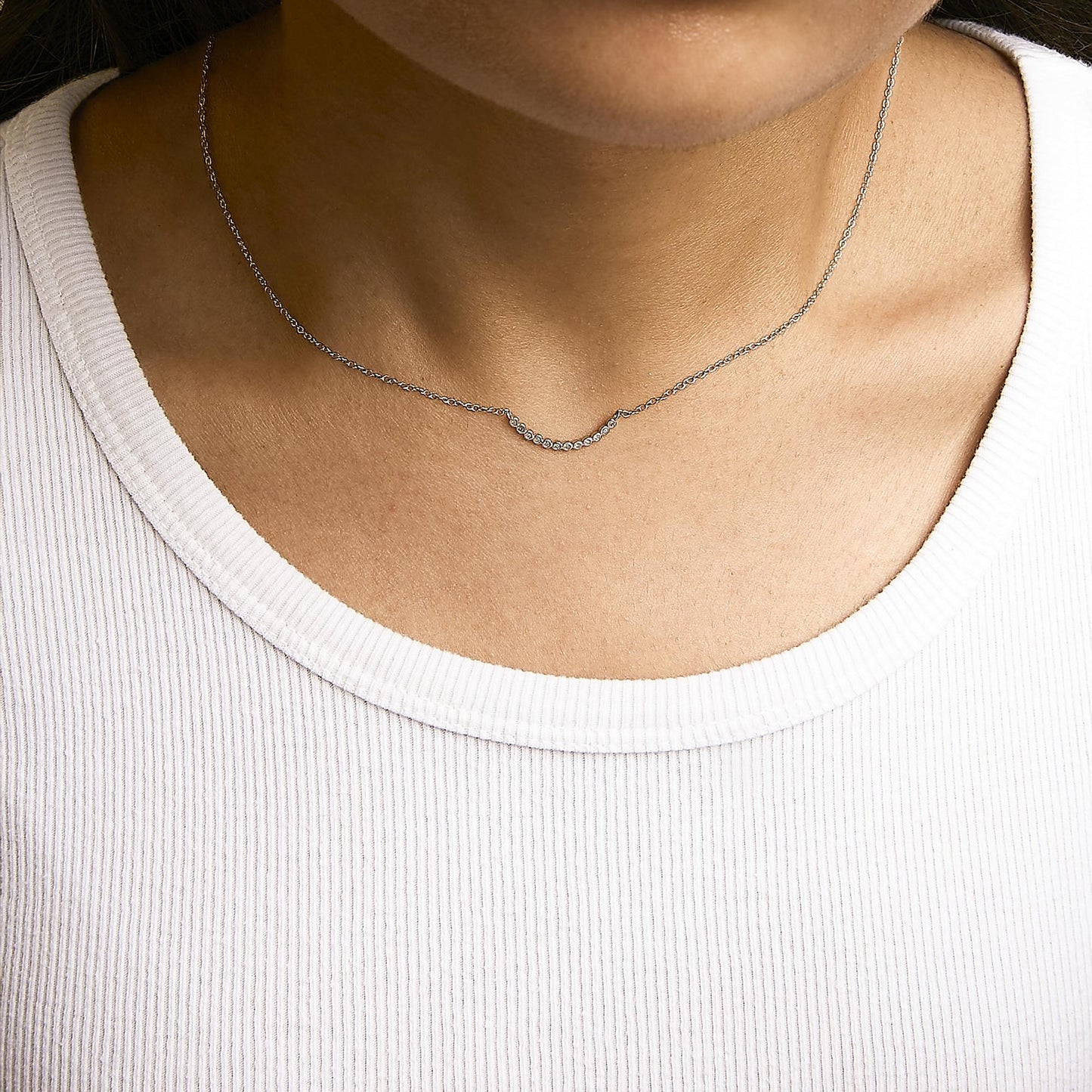 Close-up front view of a model wearing a white metal necklace with a curved bar pendant set with small round white gemstones in prong settings.