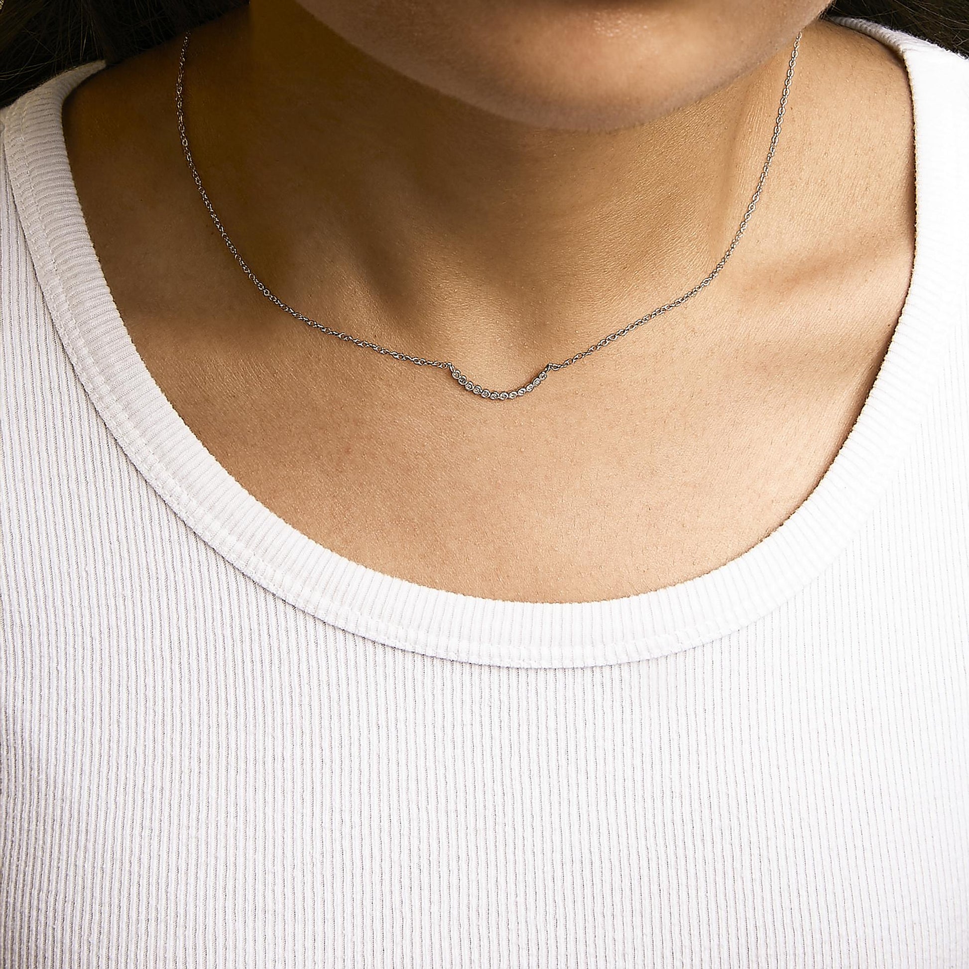 Close-up front view of a model wearing a white metal necklace with a curved bar pendant set with small round white gemstones in prong settings.