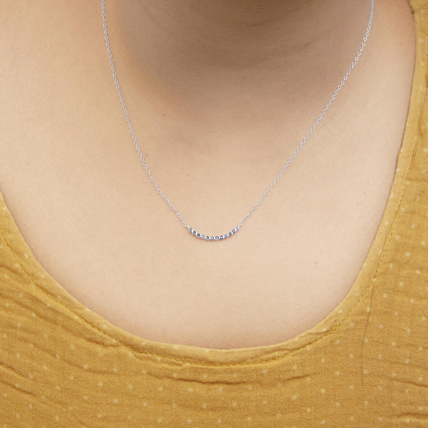 Close-up front view of a white metal necklace with a curved bar pendant set with round blue gemstones, shown worn on a model.