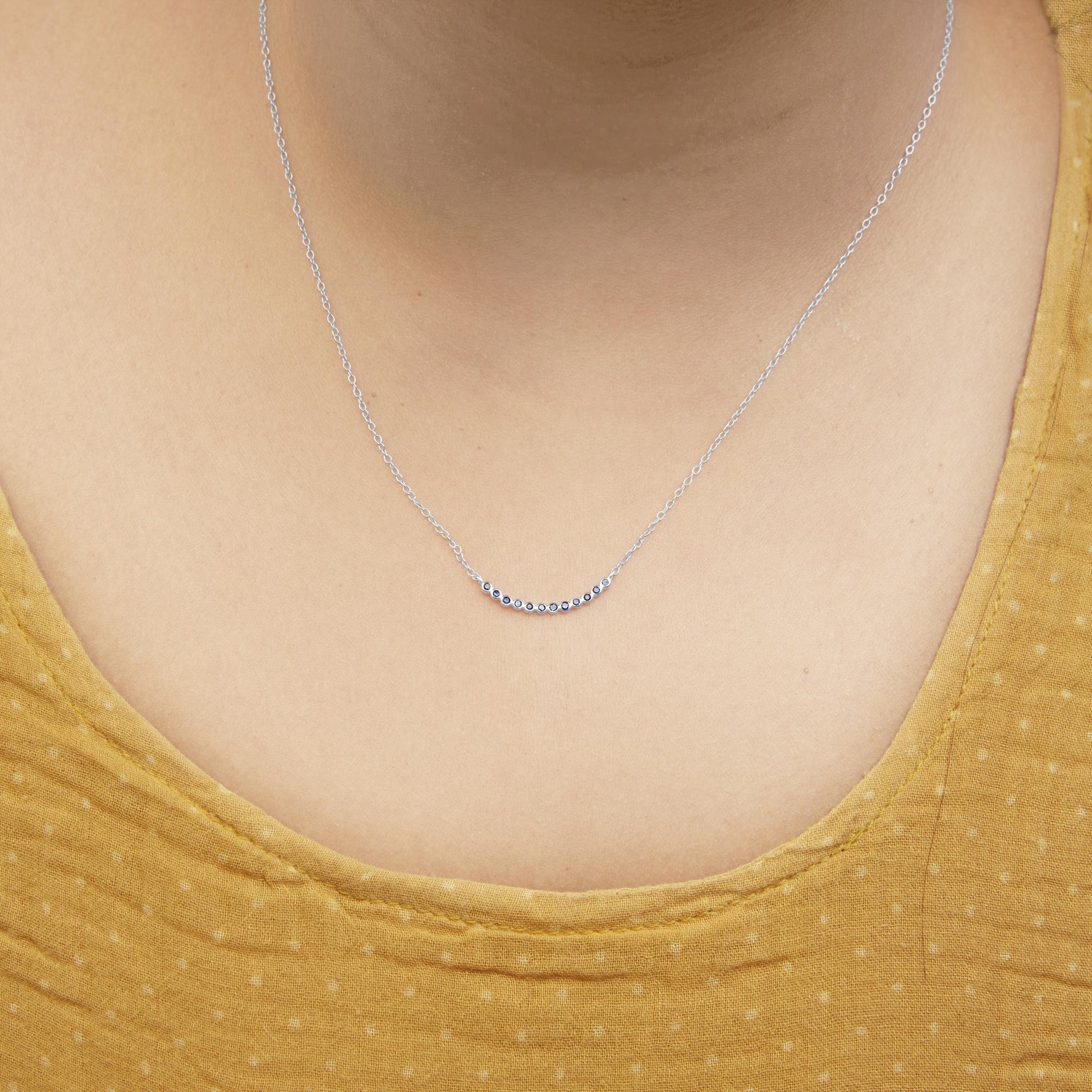 Close-up front view of a white metal necklace with a curved bar pendant set with round blue gemstones, shown worn on a model.
