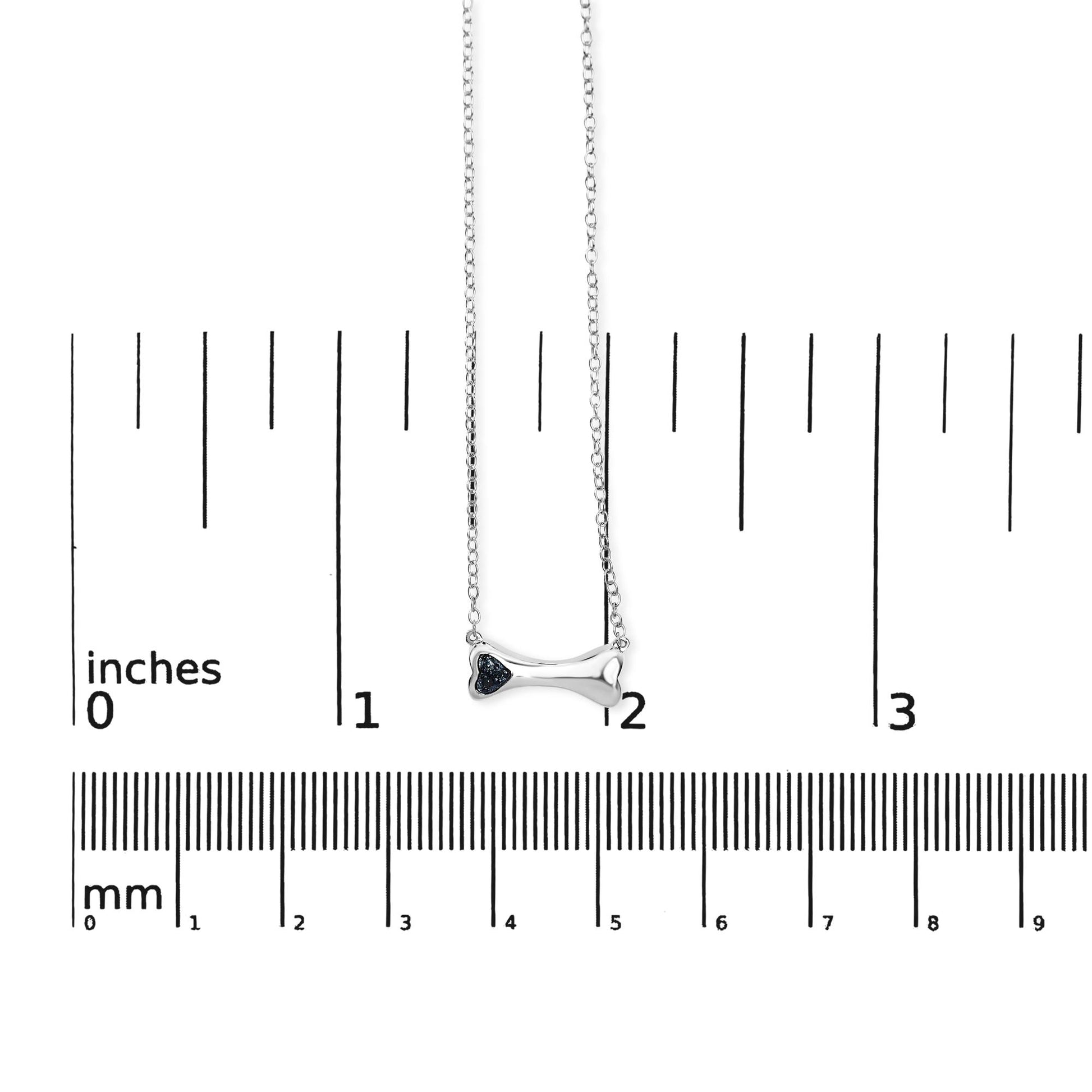 Front view of a white metal bone-shaped necklace with three blue round gemstones set in prongs, shown on a ruler for size reference.