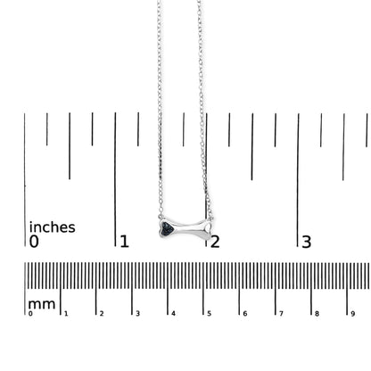Front view of a white metal bone-shaped necklace with three blue round gemstones set in prongs, shown on a ruler for size reference.