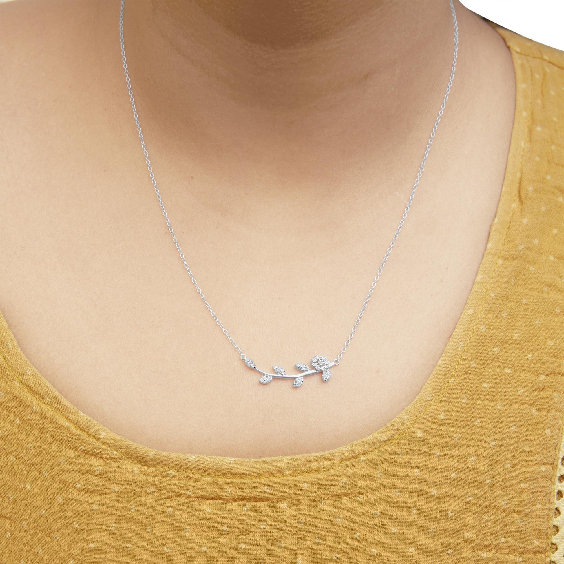 Close-up front view of a white metal necklace with a branch-shaped pendant featuring round white gemstones in prong settings, worn on a model with a mustard yellow top.