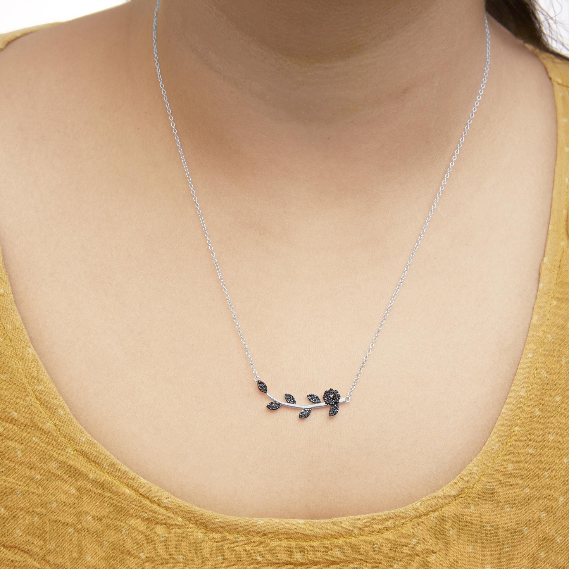 Close-up front view of a white metal necklace worn on a model, featuring a horizontal branch design with small leaves and a flower adorned with blue round prong-set gemstones.