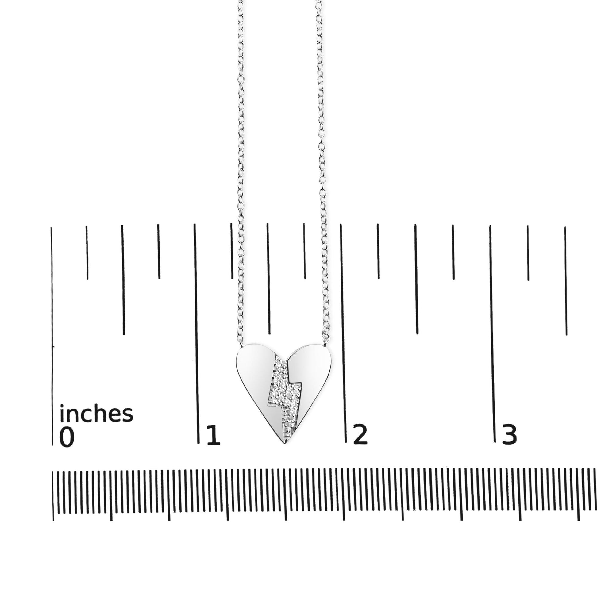 Close-up front view of white metal heart-shaped necklace pendant with a lightning bolt design set with round white gemstones, displayed on a ruler for size reference.