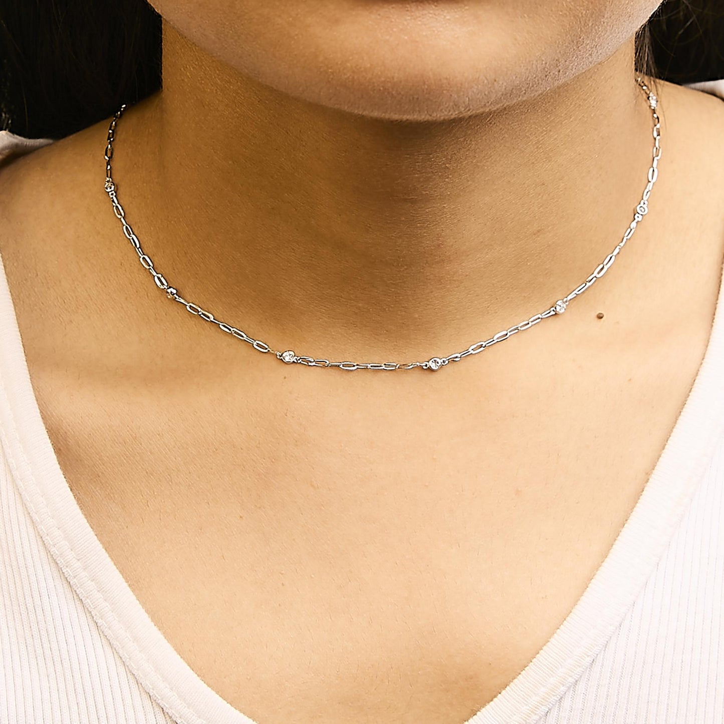 Close-up front view of a white metal necklace with small round bezel-set white gemstones, shown worn on a model.