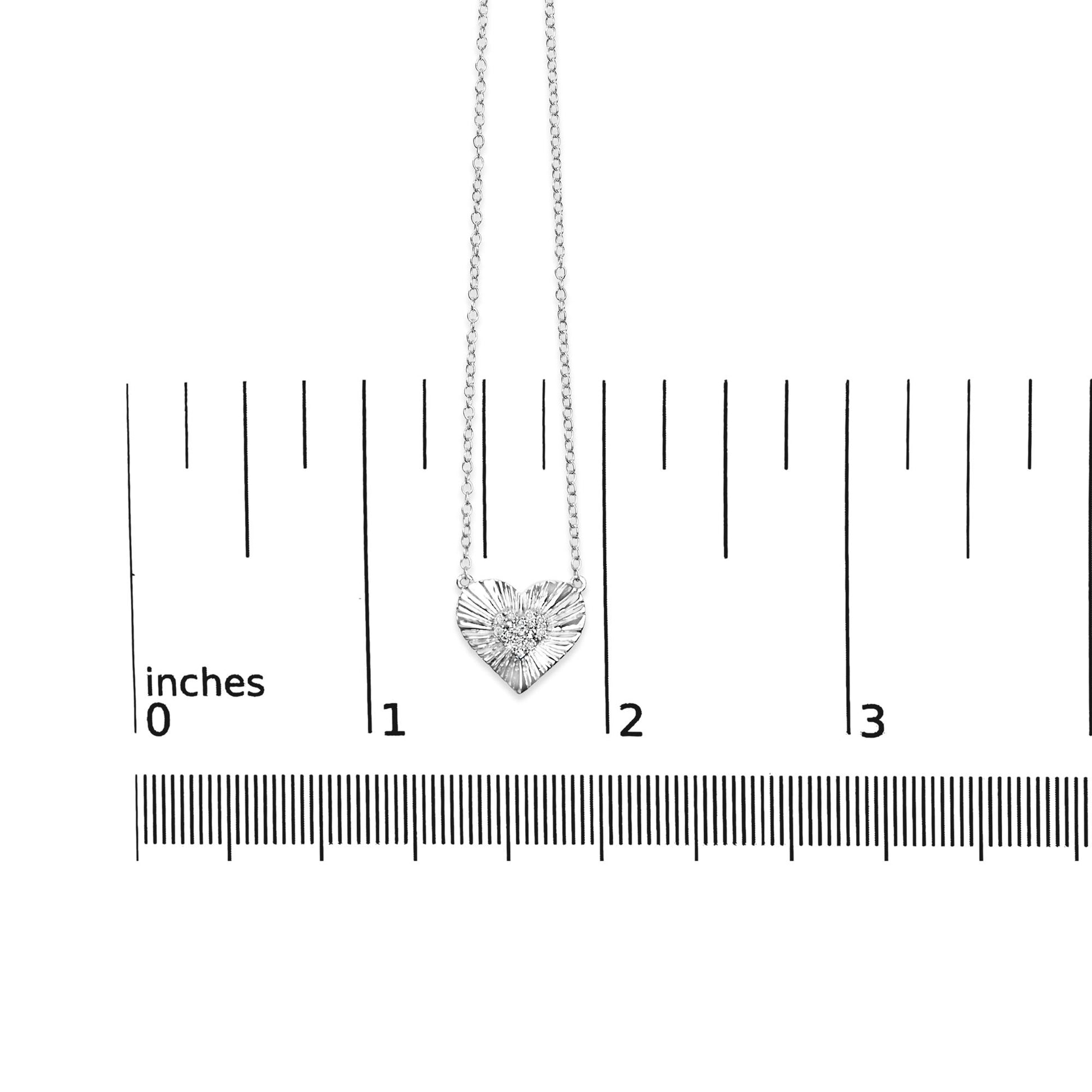 White metal heart-shaped necklace with round white gemstones in prong setting, displayed flat on a ruler for size reference, front-facing angle.