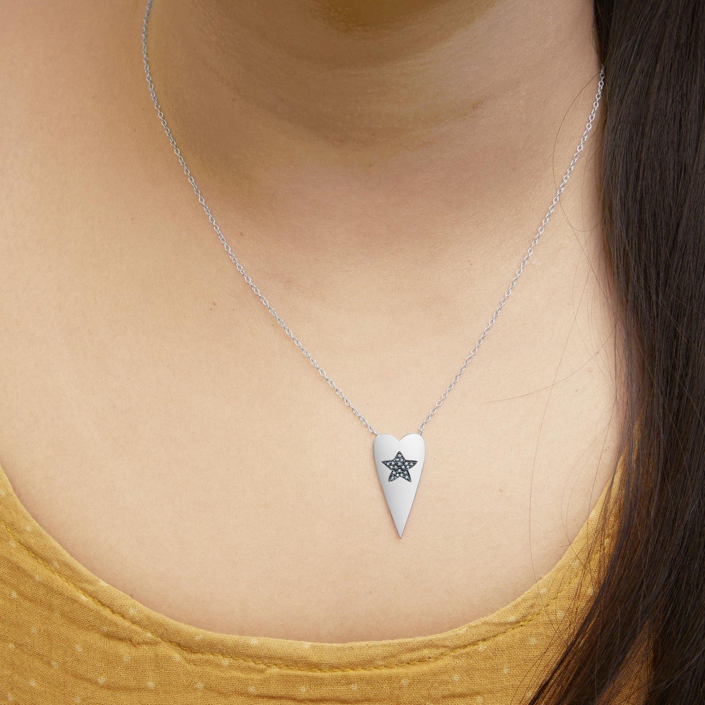 Close-up front view of a model wearing a white metal necklace with a long heart-shaped pendant featuring a star design with round blue gemstones in prong settings.