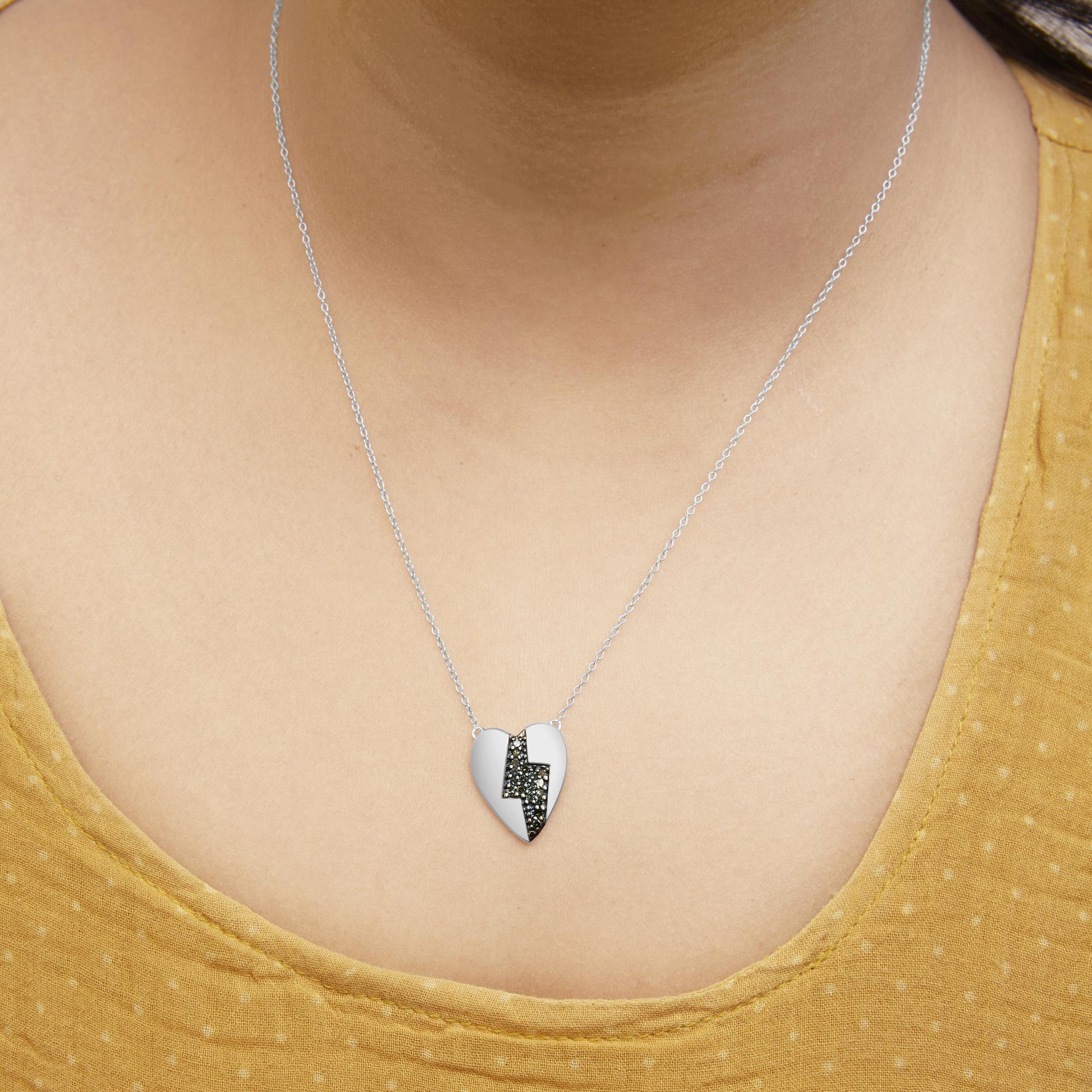 Close-up front view of a white metal heart-shaped necklace pendant with a lightning bolt design set with round blue gemstones, worn on a model.