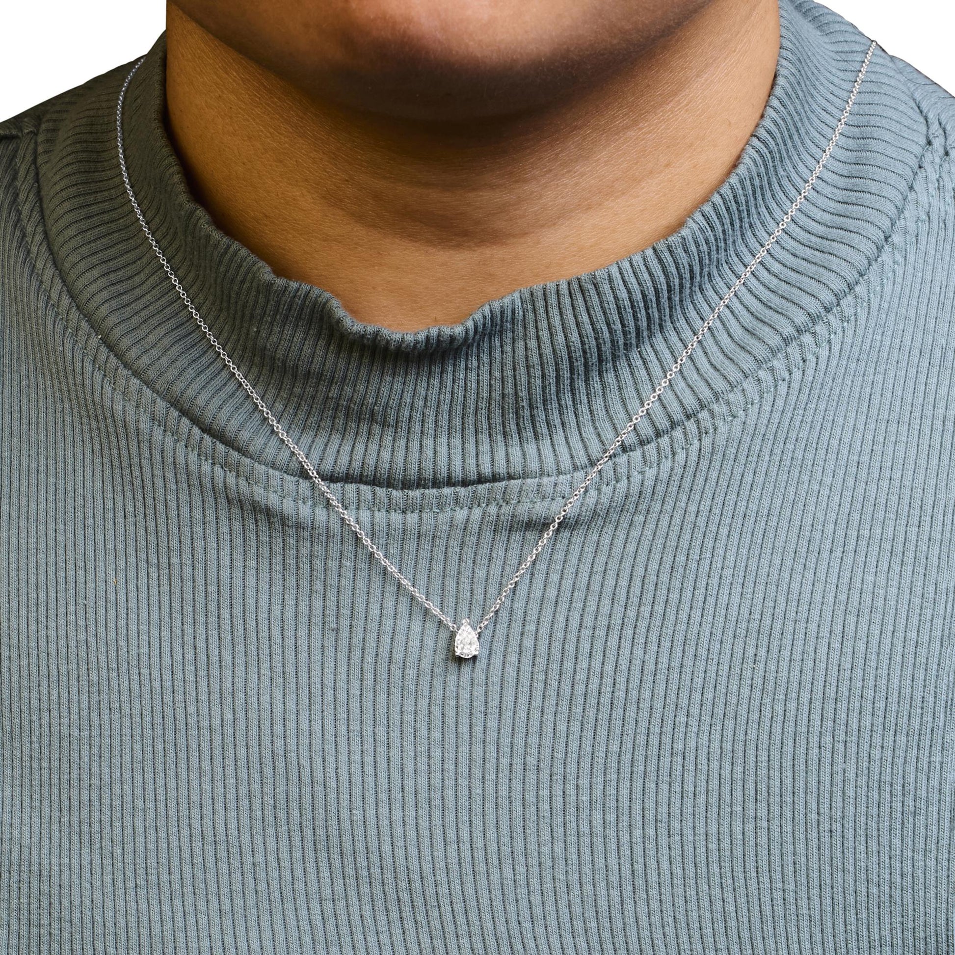 Close-up front view of a white metal necklace with a pear-shaped white gemstone pendant on a model wearing a light blue ribbed top.