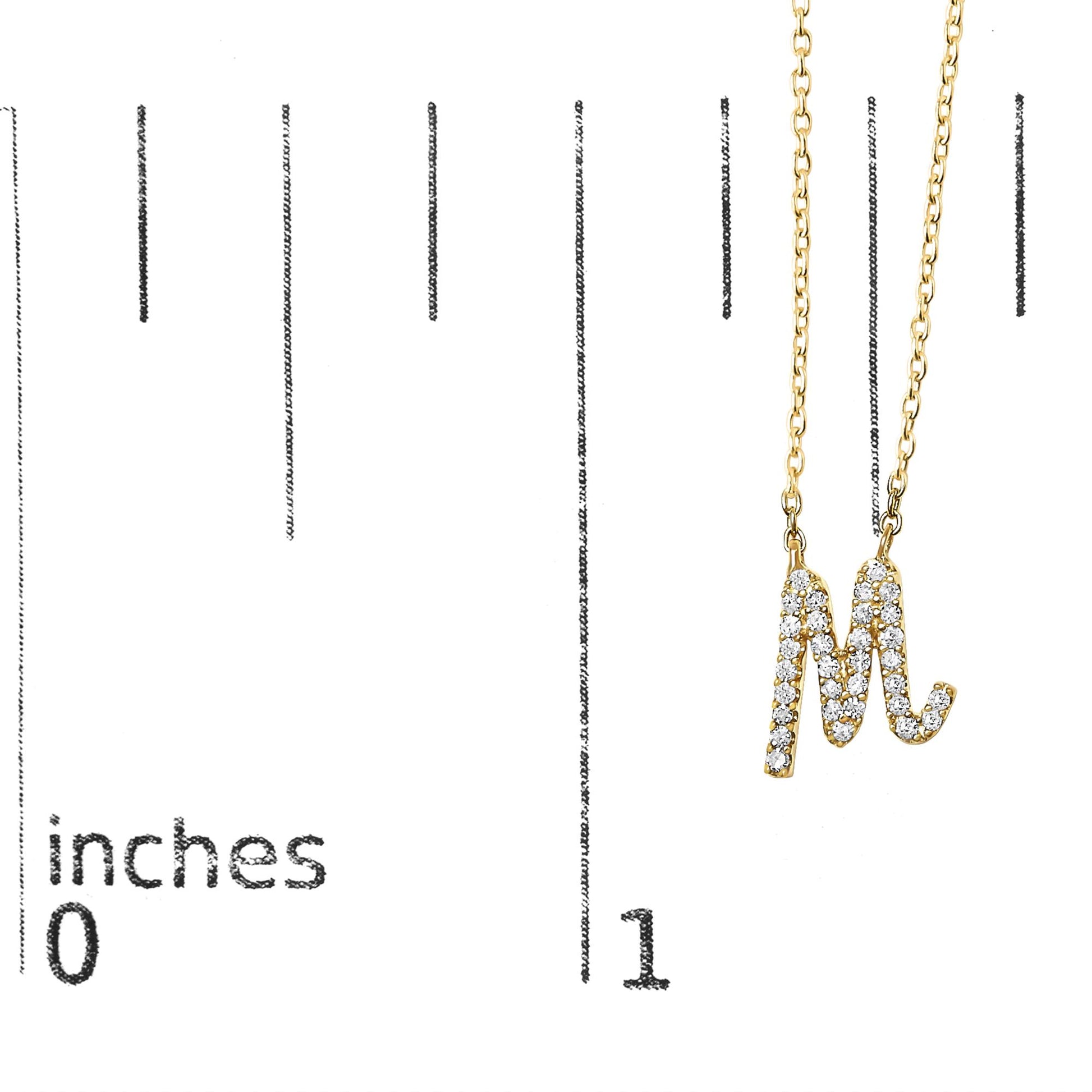 Close-up front angle of a yellow metal necklace pendant shaped like the letter M, encrusted with round white gemstones in a micro-pave setting, shown on a scale.