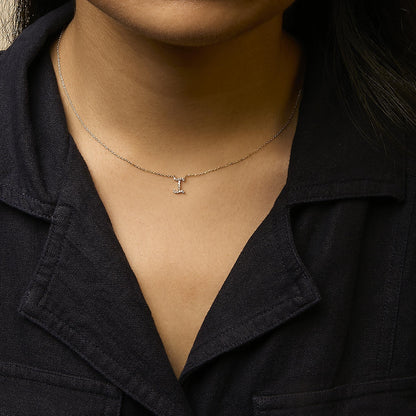 Close-up front view of a model wearing a yellow metal necklace with a small, vertically oriented pendant shaped like the letter 'I,' featuring white round gemstones in a micro-pave setting.