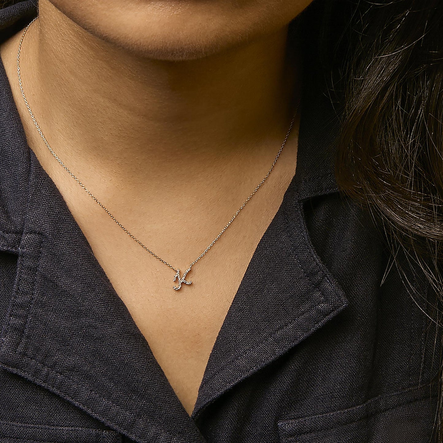Close-up front view of a model wearing a yellow metal necklace with a pendant shaped like the letter 'K' adorned with small white gemstones in a micro-pave setting.