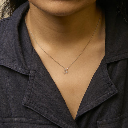 Close-up front view of a model wearing a yellow metal necklace with a pendant shaped like the letter 'M' encrusted with small white gemstones in a micro-pave setting.
