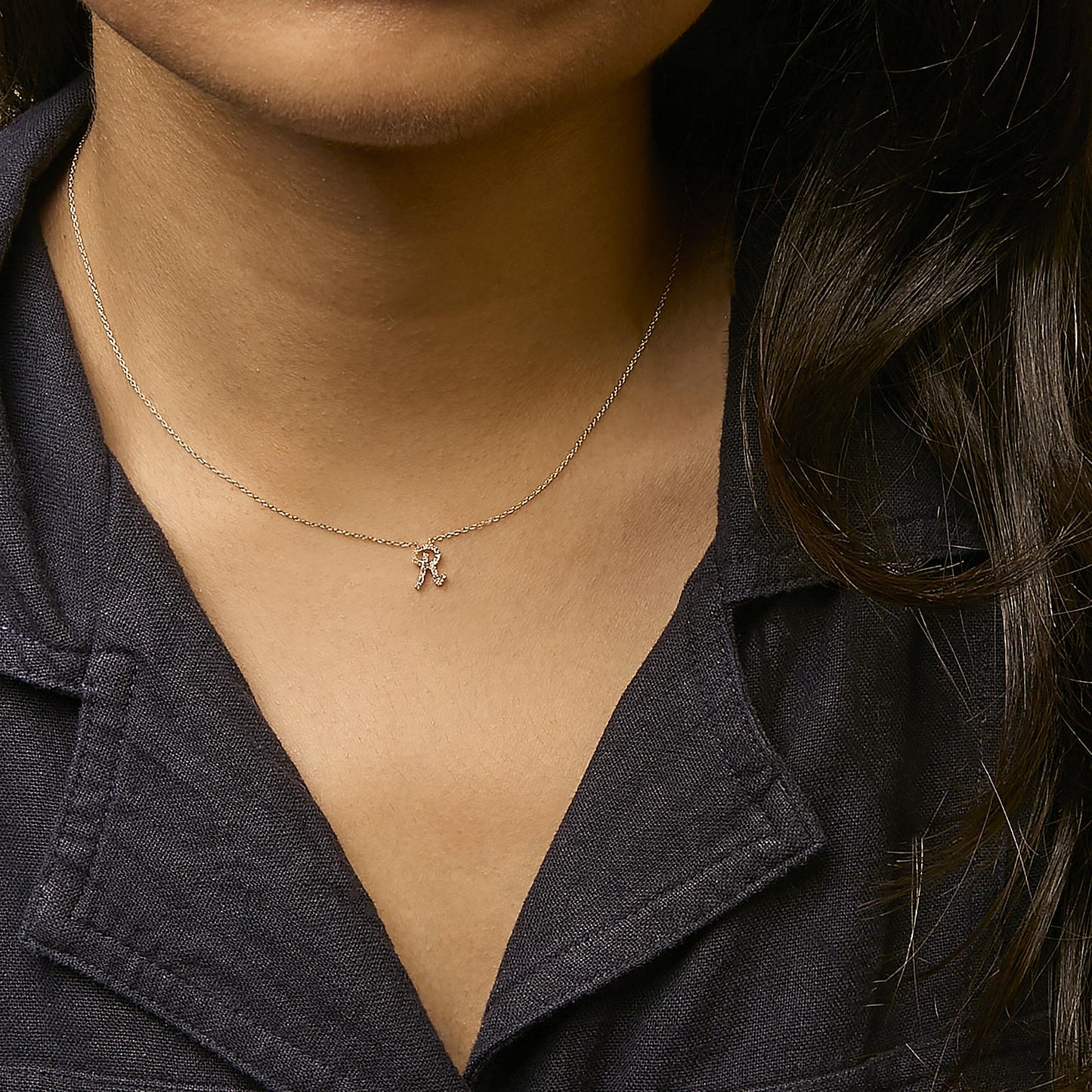 Close-up front view of a model wearing a yellow metal necklace with a small pendant shaped like the letter 'R' adorned with tiny white gemstones in a micro-pave setting.