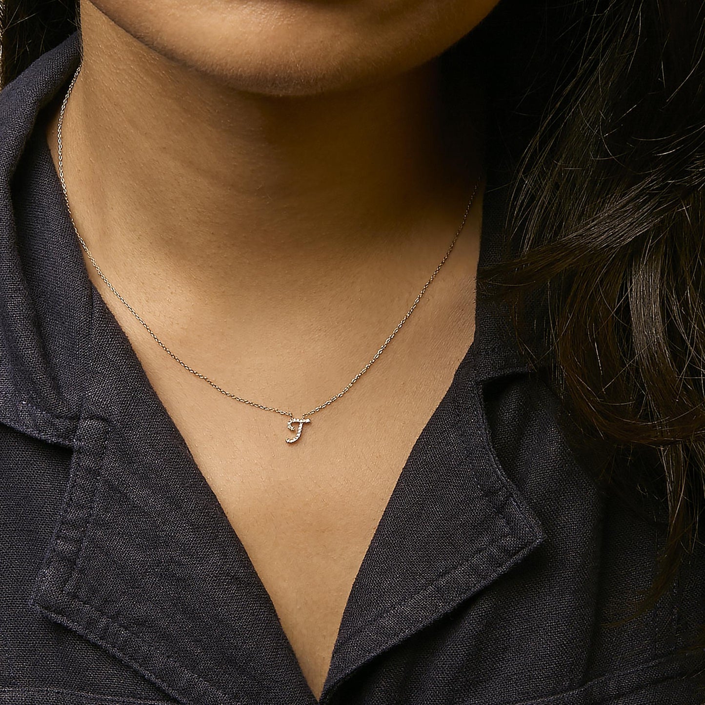 Yellow metal necklace with a pendant shaped like the letter 'J' featuring white round gemstones in a micro-pave setting, shown worn on a model from a front angle.