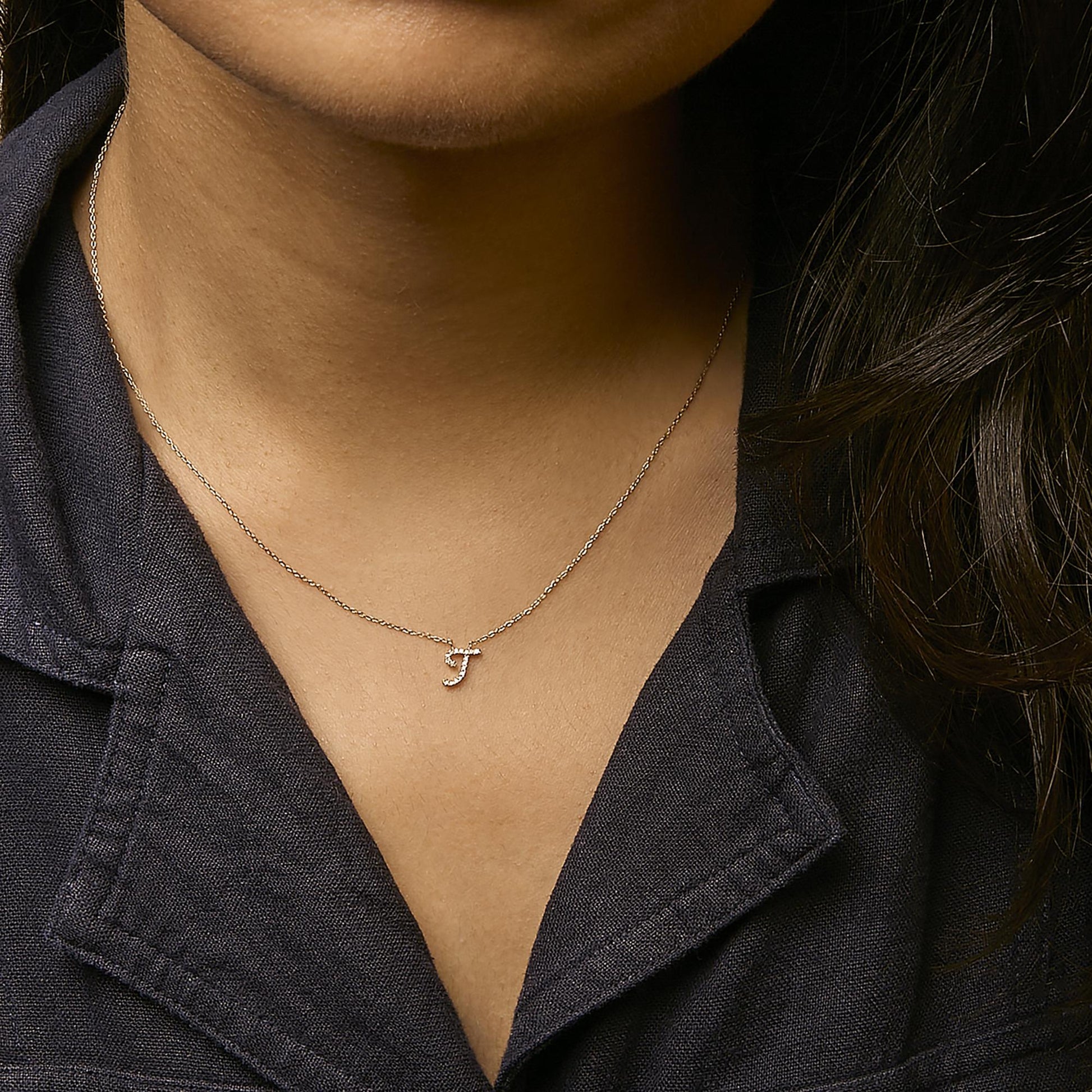Yellow metal necklace with a pendant shaped like the letter 'J' featuring white round gemstones in a micro-pave setting, shown worn on a model from a front angle.