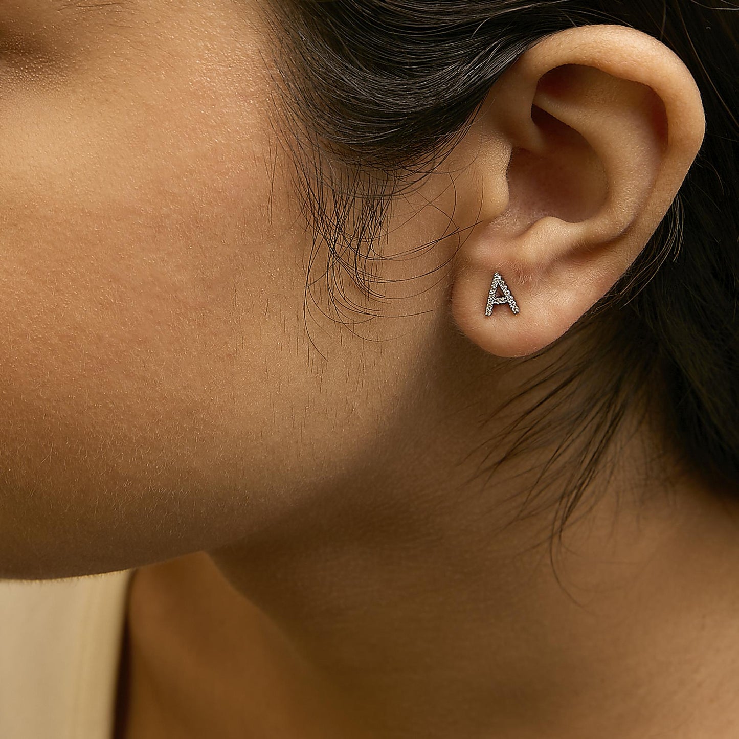 Close-up side view of a model wearing a white metal earring shaped like the letter 'A' with micro-pave set round white gemstones.