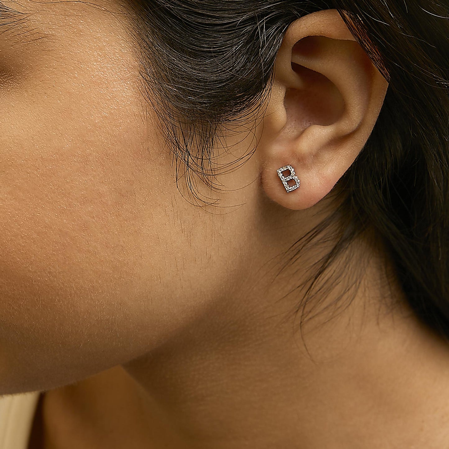 Close-up side view of a model wearing a white metal earring shaped like the letter 'B' adorned with round white gemstones in a micro-pave setting.
