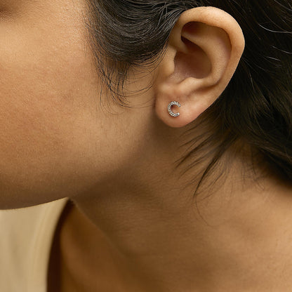 Close-up side view of a model wearing small white metal crescent-shaped earrings with white round gemstones in a micro-pave setting.