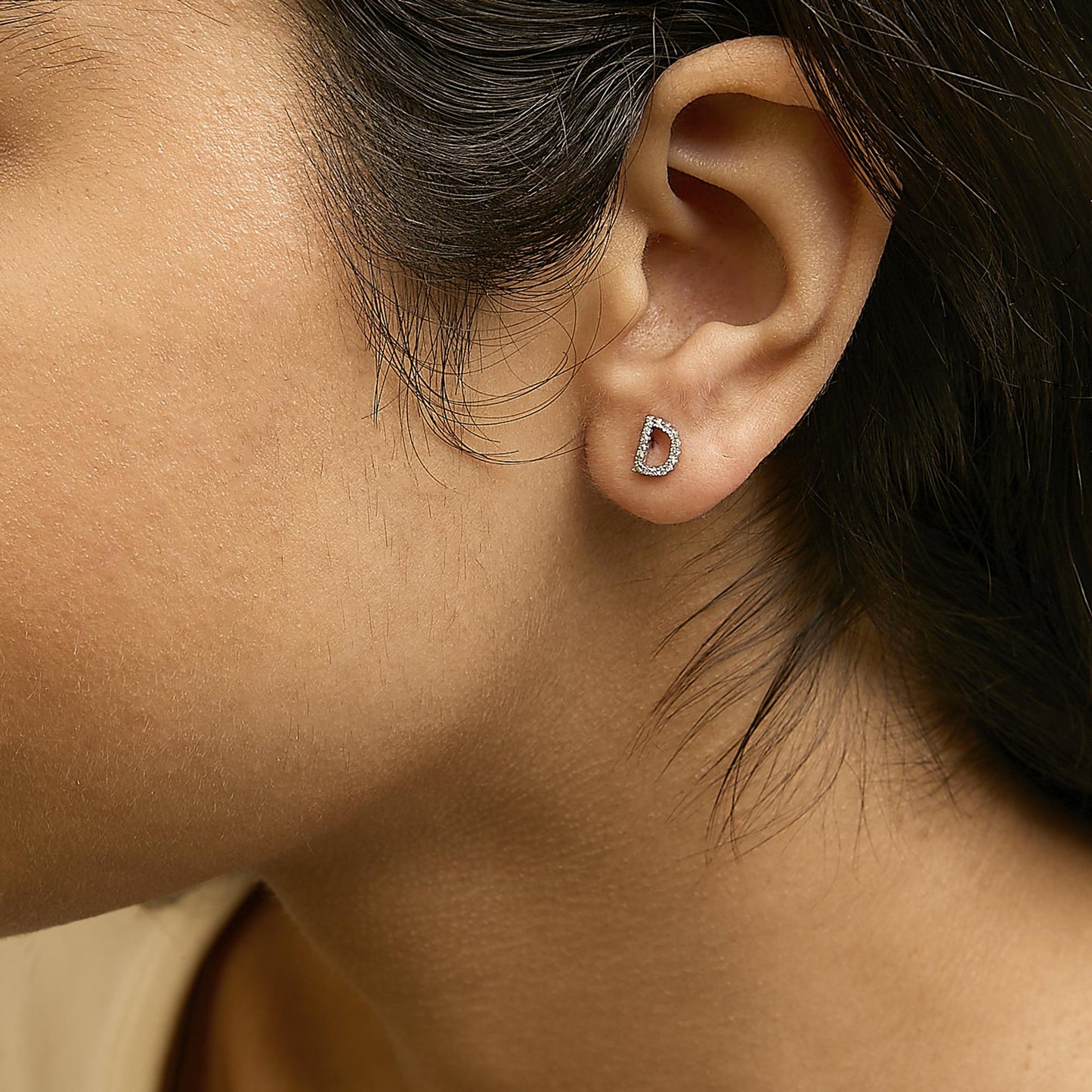 Close-up side view of a model wearing a white metal earring shaped like the letter D, encrusted with small round white gemstones in a micro-pave setting.