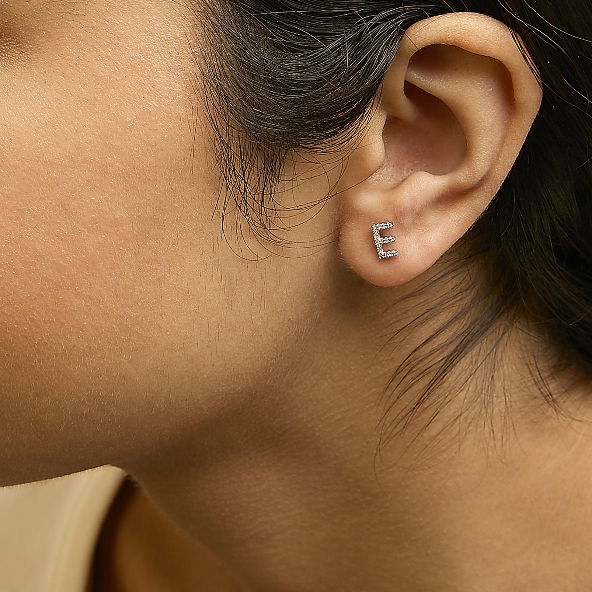Close-up side view of a model wearing a white metal stud earring shaped like the letter 'E' adorned with small white gemstones in a micro-pave setting.