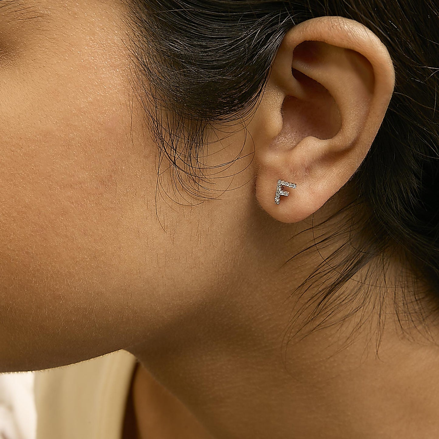 Close-up side view of a model wearing a white metal earring shaped as the letter F, adorned with small round white gemstones in micro-pave setting.