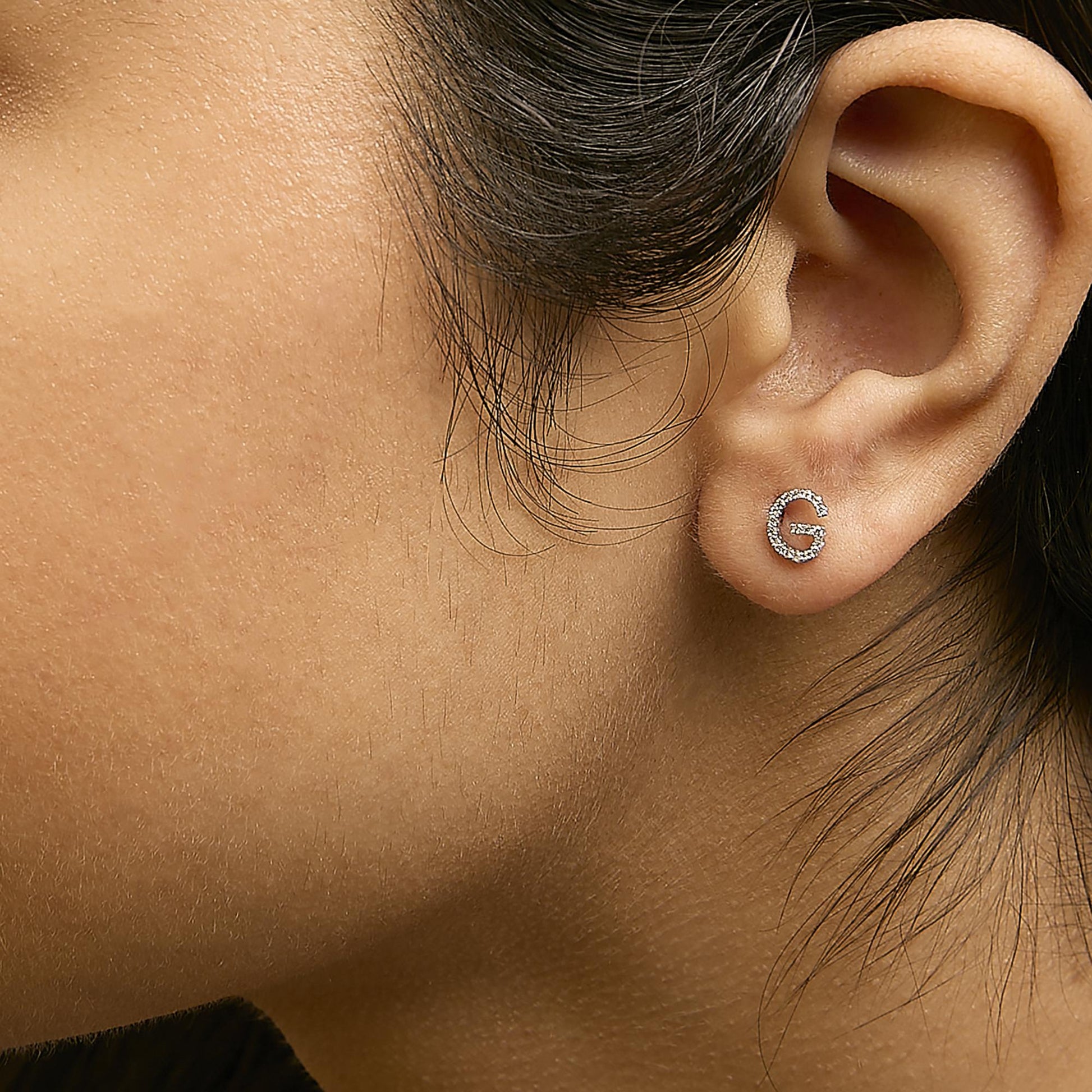 Close-up side view of a model wearing a white metal earring shaped like the letter 'G' adorned with small round white gemstones in a micro-pave setting.