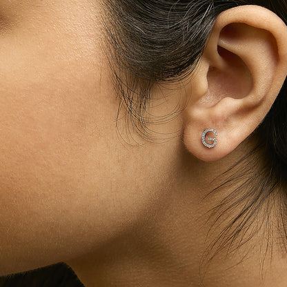 Close-up side view of a model wearing a white metal earring shaped like the letter 'G' adorned with small round white gemstones in a micro-pave setting.